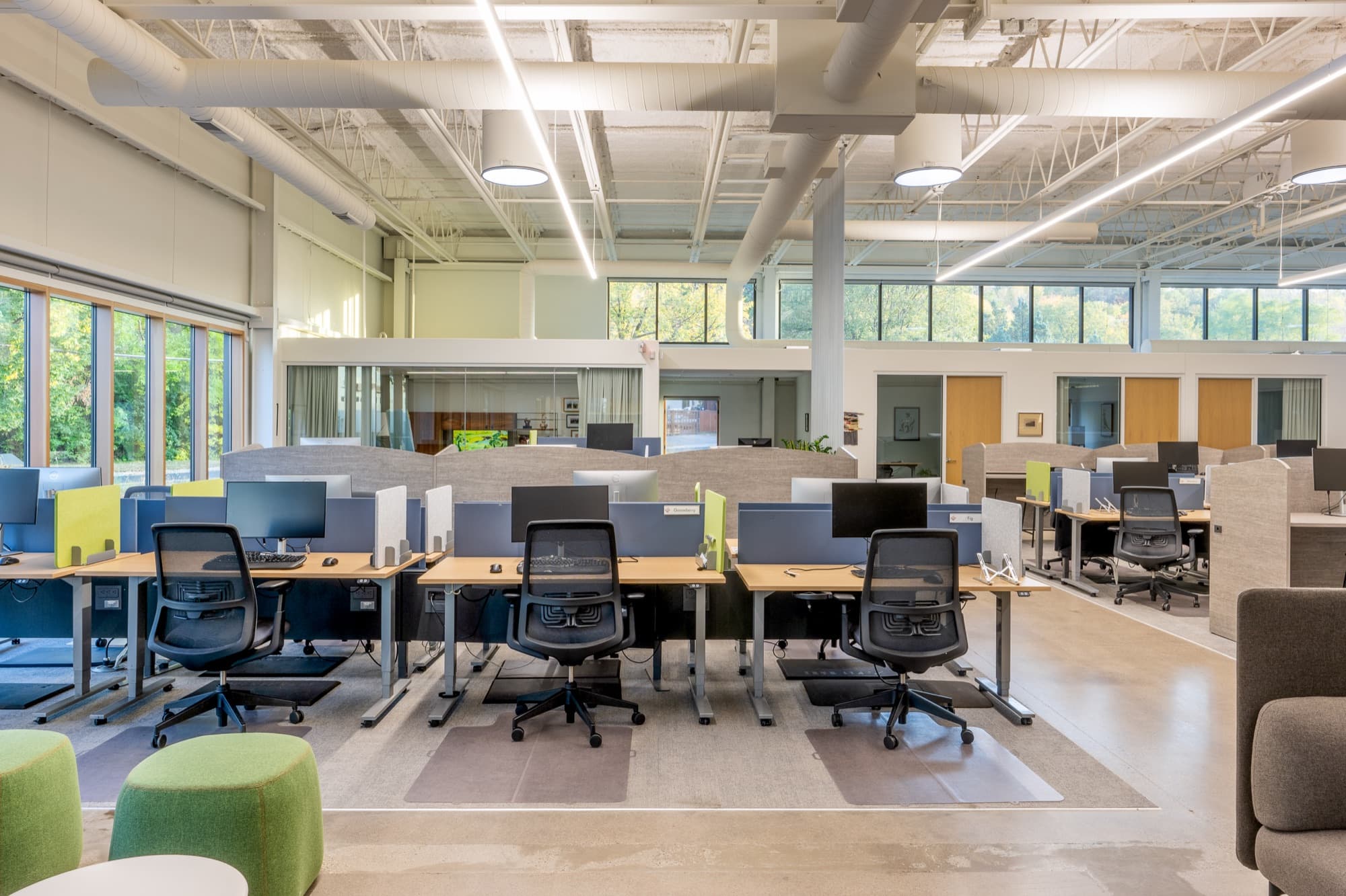 Open office with rows of desks and green stools beside tall daylight windows